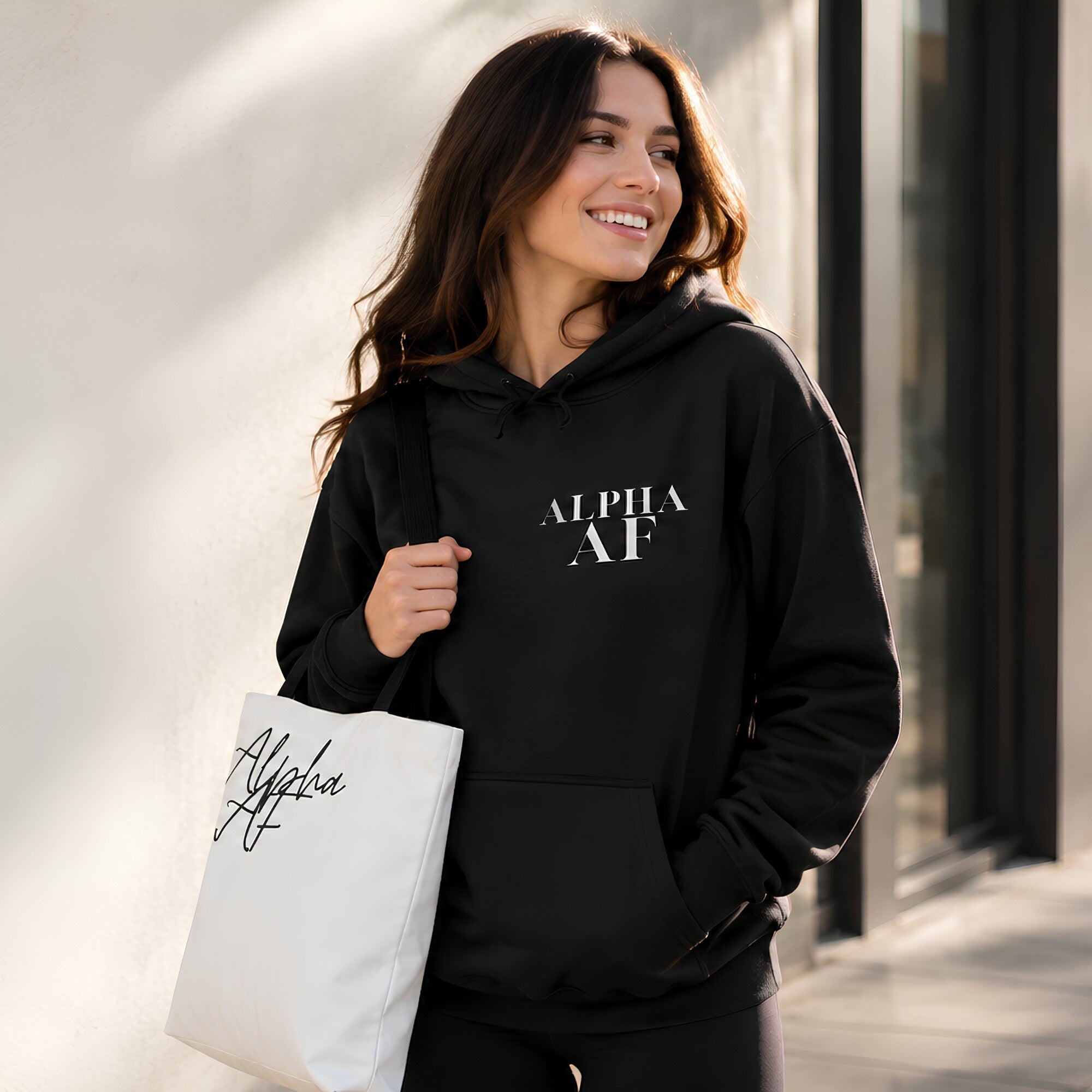 A woman wearing a black hoodie with 'ALPHA AF' printed in white on the chest, carrying a white tote bag with black script, standing against a sunlit beige wall.